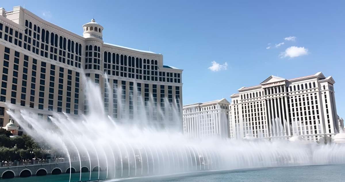 The Bellagio Fountains in Las Vegas spraying water high into the air with Caesars Palace in the background on a clear, sunny day.