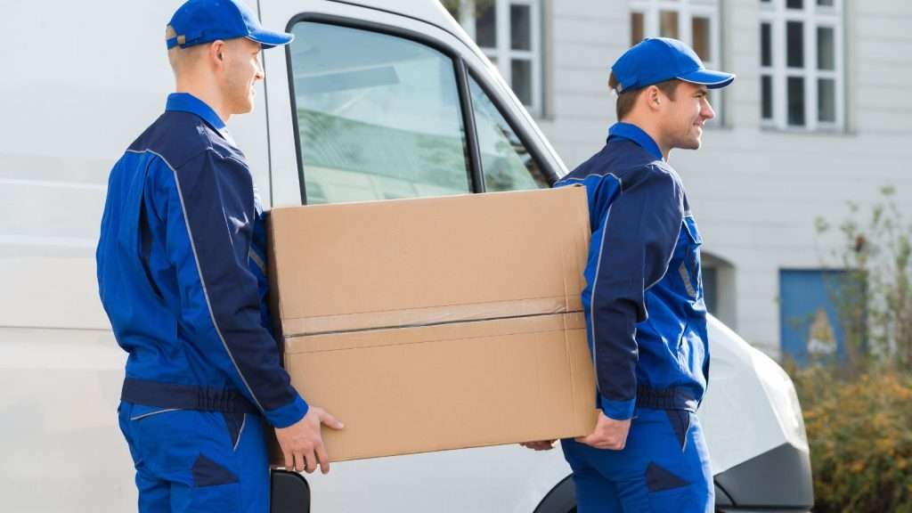 Two professional movers in blue uniforms carrying a large cardboard box beside a moving truck during a residential move.