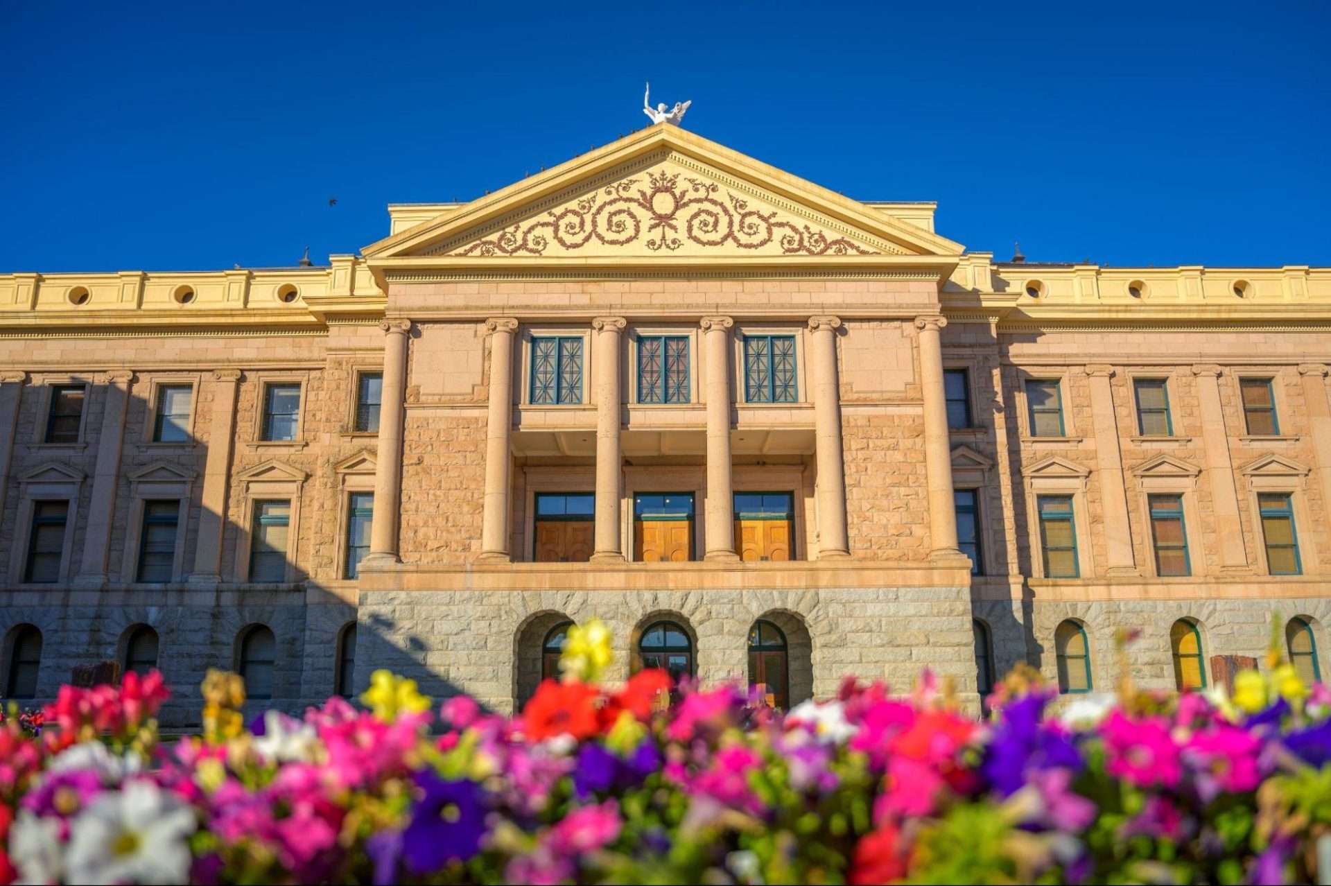 Arizona State Capitol in Phoenix, AZ, with colorful flowers in the foreground under a blue sky.