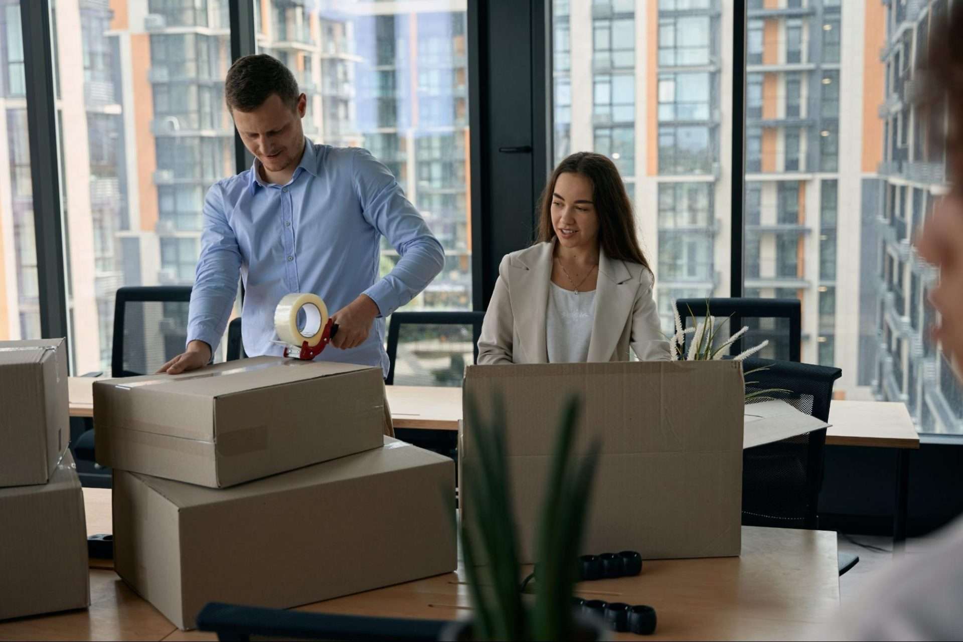 Two employees, one male, and one female, stand behind desk taping up moving boxes.