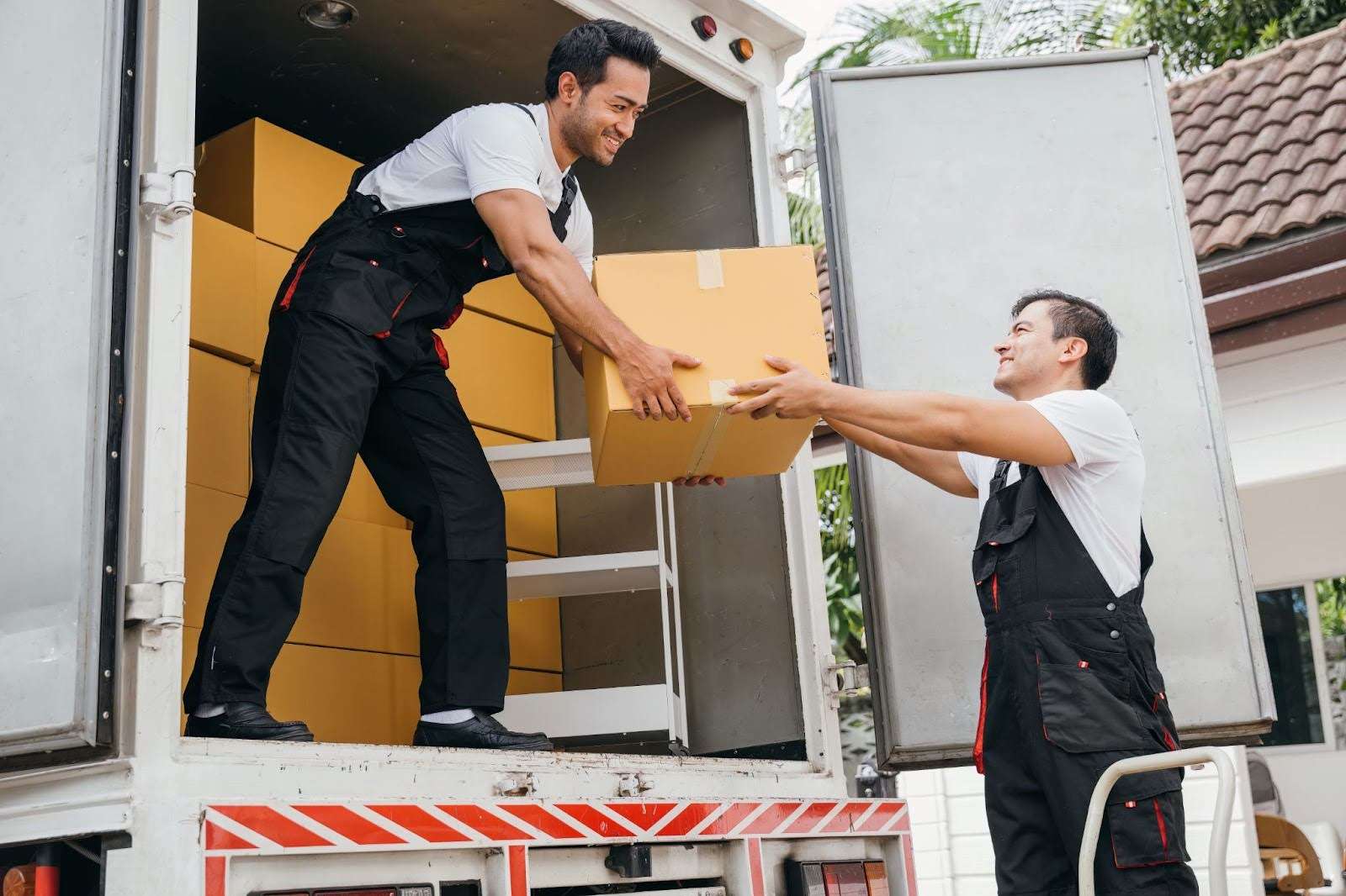 One mover standing in the back of a moving truck handing a box to another mover on the ground.