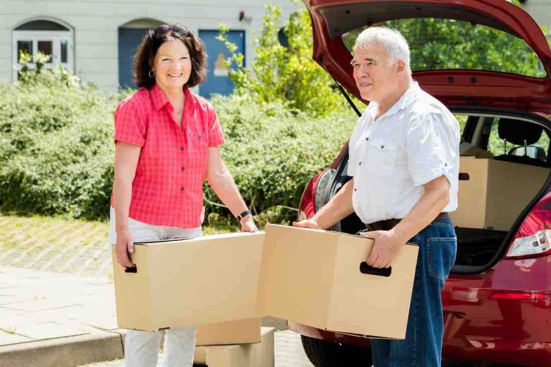 Two older moving customers holding boxes in front of an open trunk