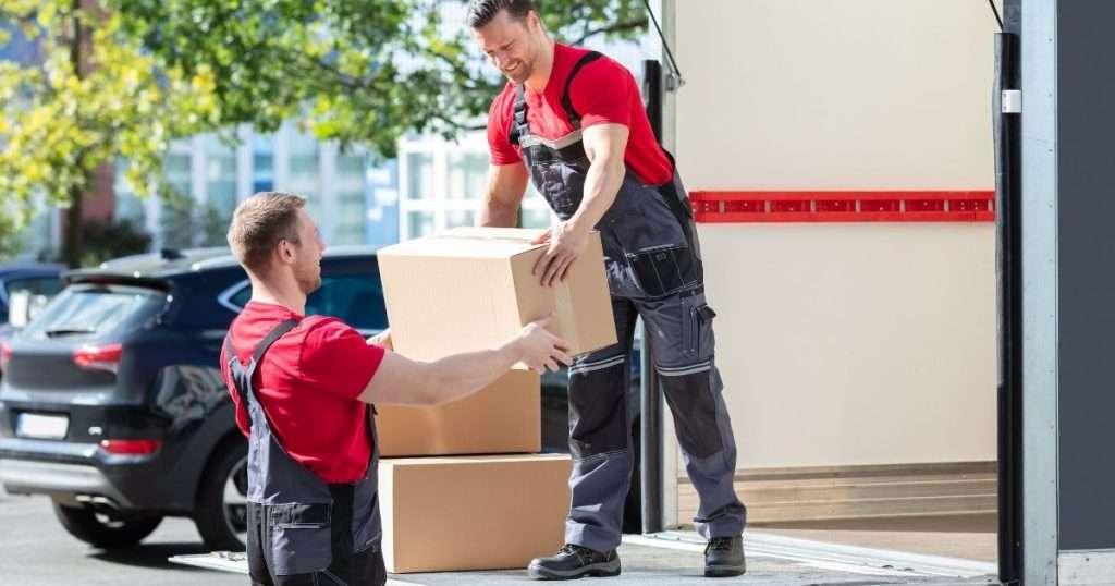 Two professional Surprise movers in red shirts and work overalls loading cardboard boxes onto a moving truck on a sunny day.