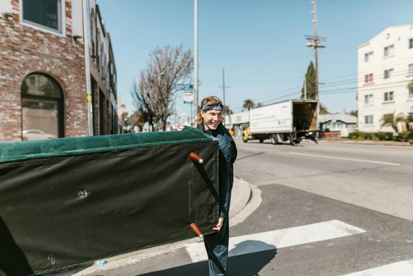 Smiling man carrying a couch across a crosswalk.