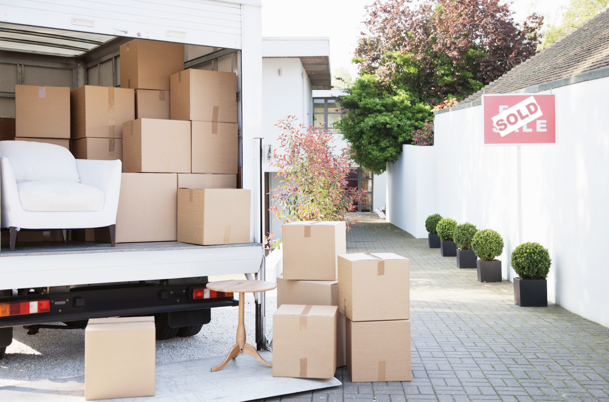 Boxes and white chair sitting in the back of a moving truck outside of a home with a 'sold' sign on it.