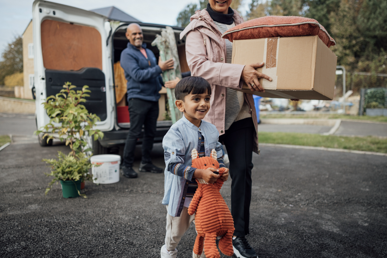 Young child holding a stuffed animal and smiling as his parents help to unload a moving van in the background.