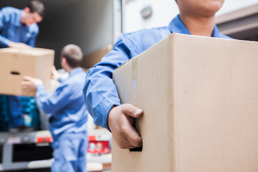 Close up of mover carrying a moving box while two other movers unload boxes from a moving truck in the background.